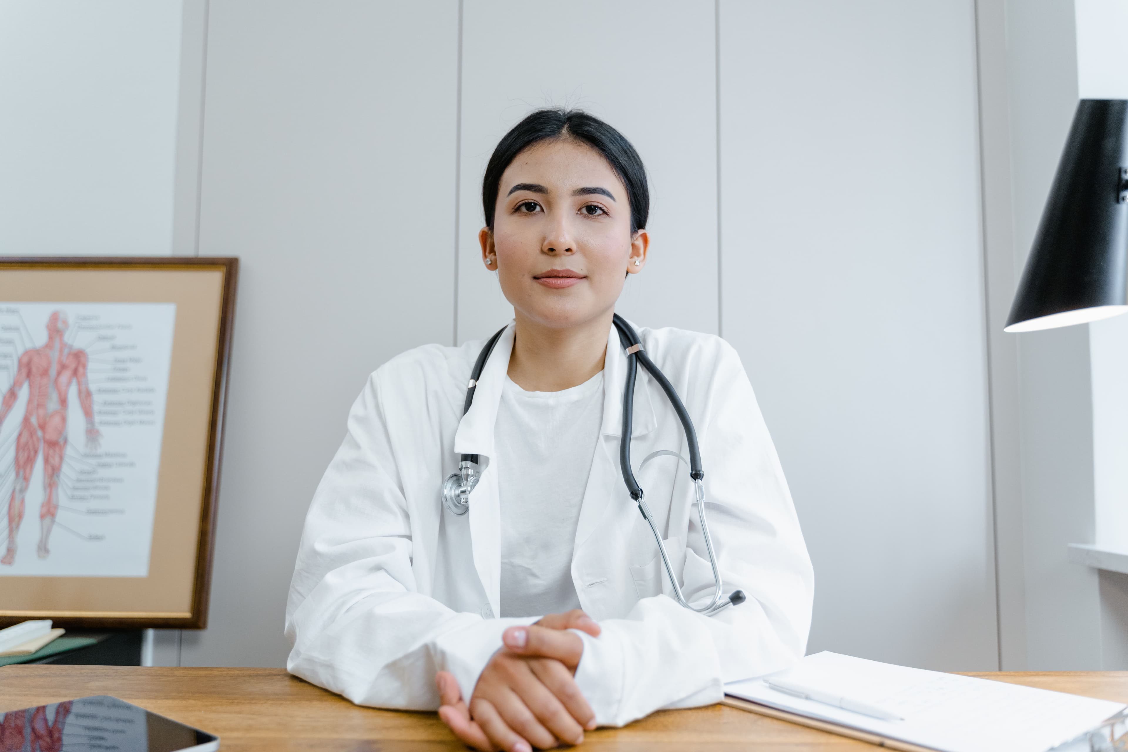 A doctor in a white coat with a stethoscope around their neck, seated at a desk with hands clasped, facing the camera